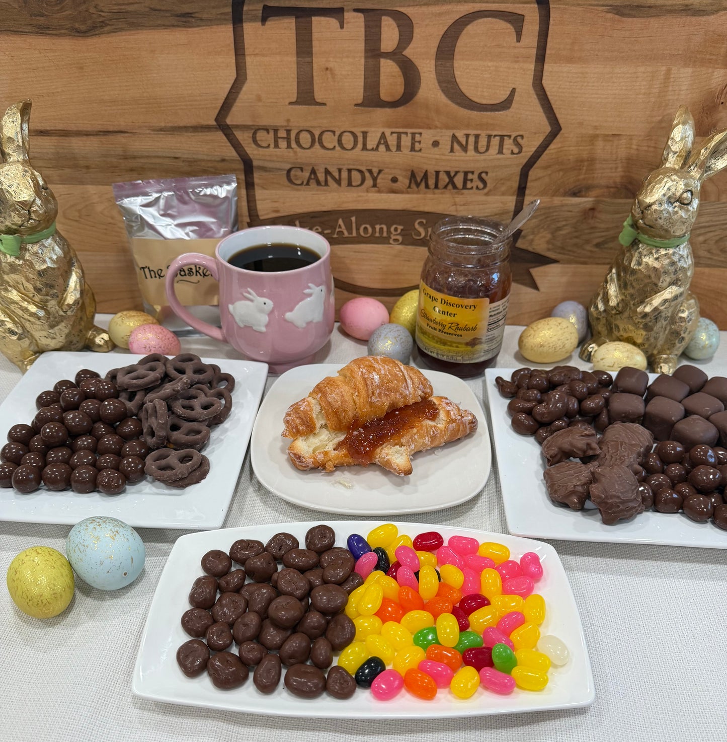 Assorted candies, a croissant with jam, and a mug on a table with 'TBC Chocolate Nuts Candy Mixes' sign in the background.