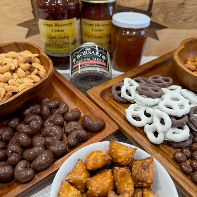 Assorted snacks including pretzels, chocolate-covered cashews, and nuts on a wooden tray with bottles in the background.