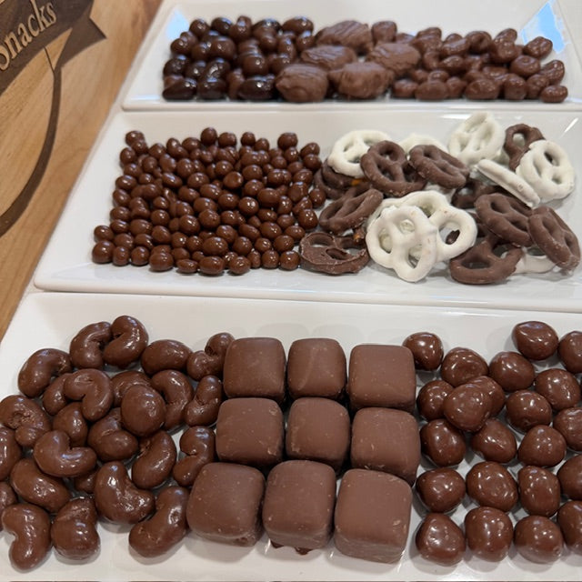 Assorted chocolate-covered snacks including nuts, pretzels, and candy on a white tray.