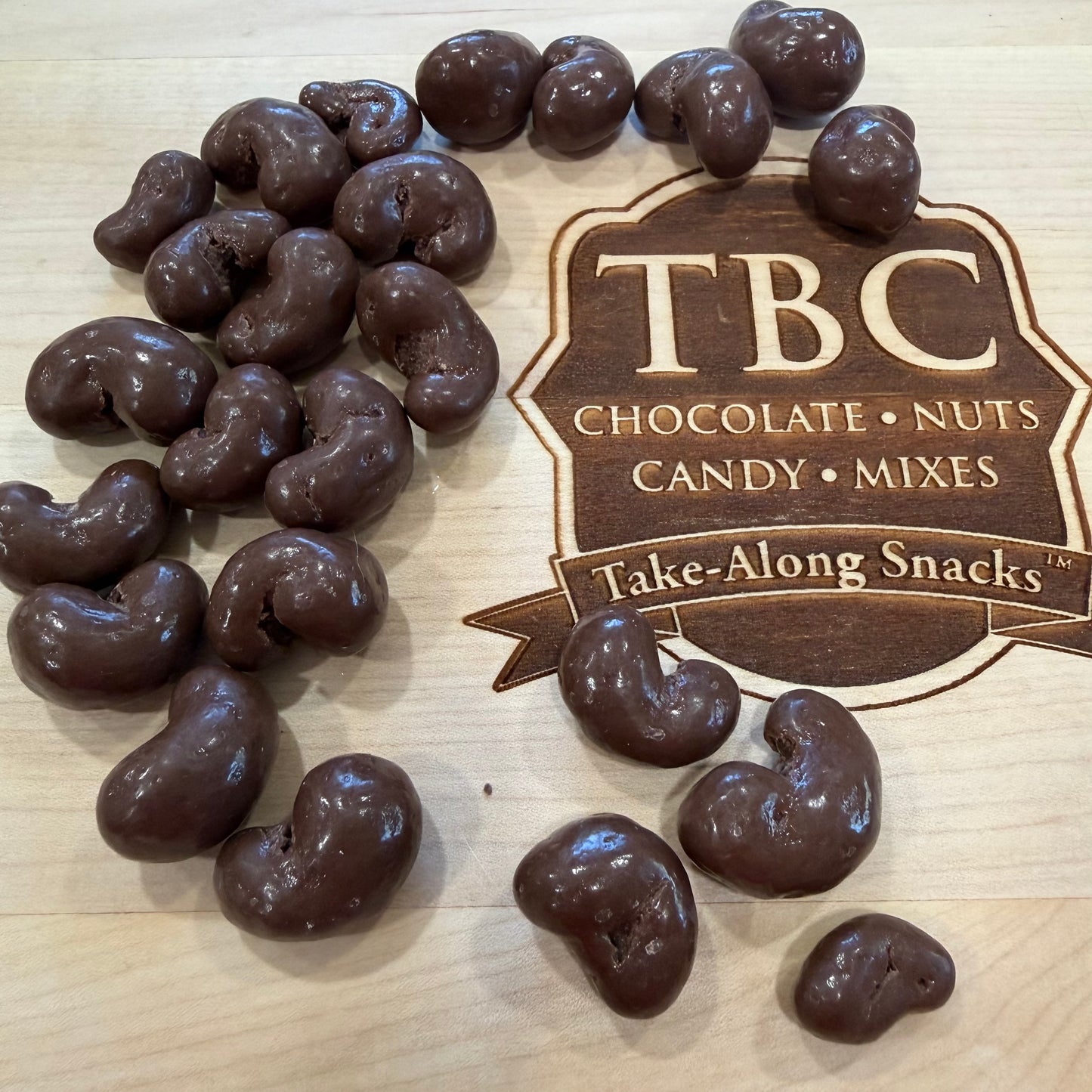 Chocolate-covered cashews on a wooden surface with TBC Chocolate Nuts Candy Mixes branding.