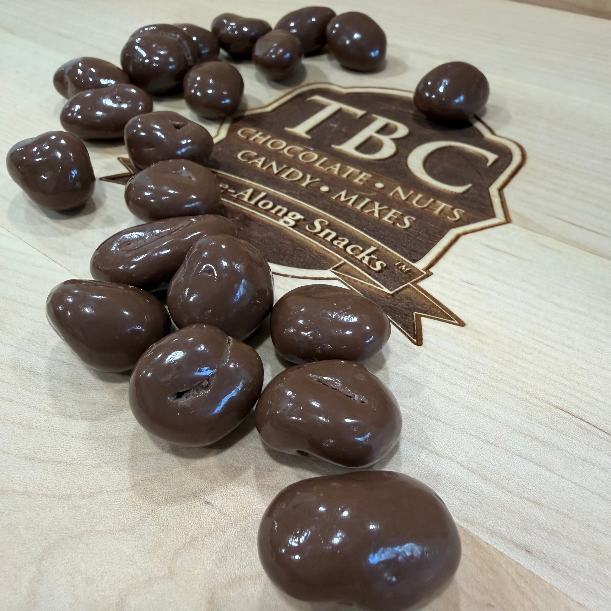 Chocolate-covered candies on a wooden surface with TBC Chocolate Candy Nuts logo.