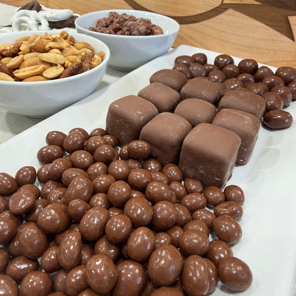 Assorted chocolate candies and nuts on a white surface with bowls of additional ingredients in the background.