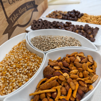 Assorted trail mix in a white divided bowl with a wooden box labeled 'Take-Along Snacks' in the background.
